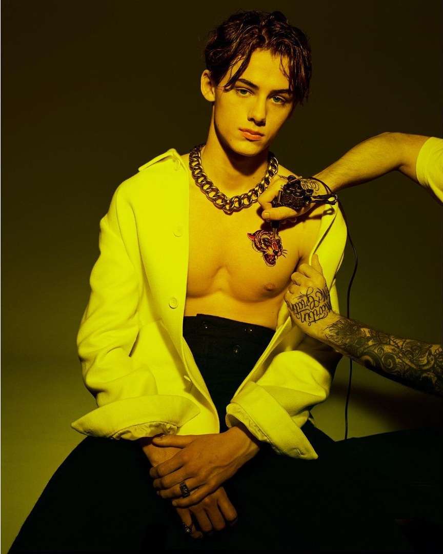 Young man in a yellow jacket, no shirt, with tiger chest tattoo. Artist adding new tattoos on hands and chest in a studio.