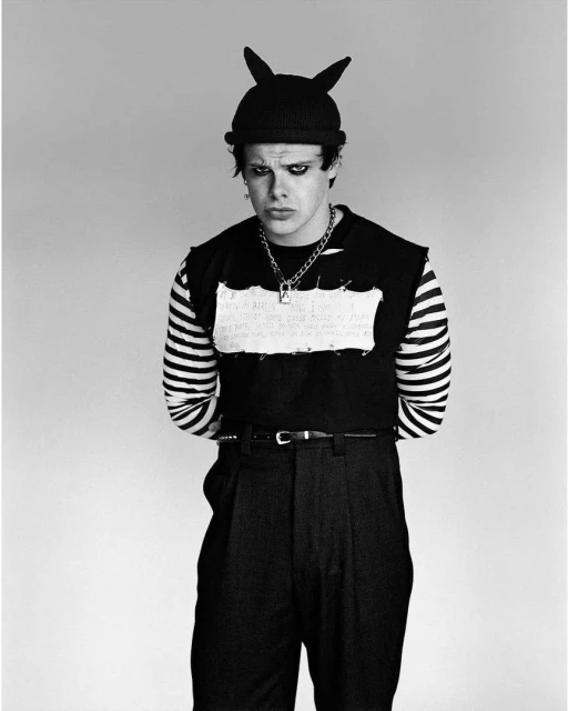 Black and white portrait of an adult male with dark clothing, a beanie with ears, and a chain necklace, standing against a light background.