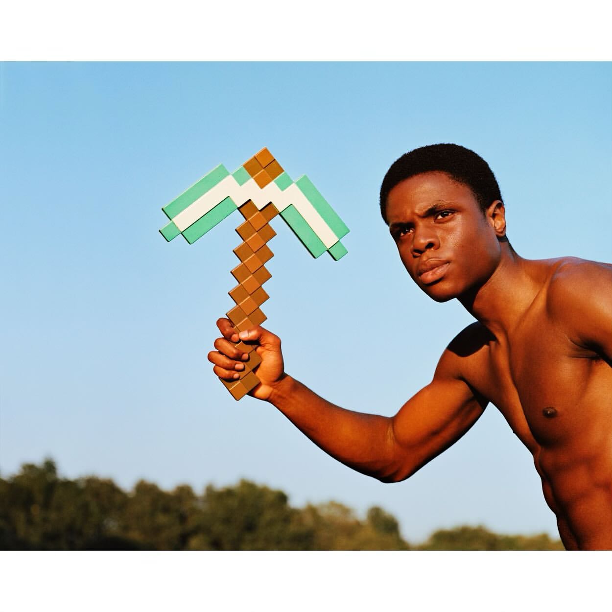 Athletic Black man holds a Minecraft-style pickaxe outdoors against a clear blue sky with green trees in the distance.