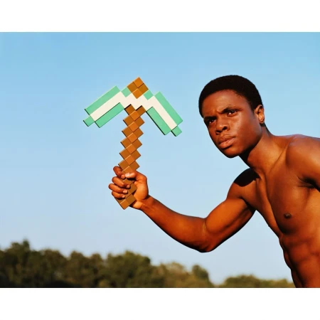 Athletic Black man holds a Minecraft-style pickaxe outdoors against a clear blue sky with green trees in the distance.
