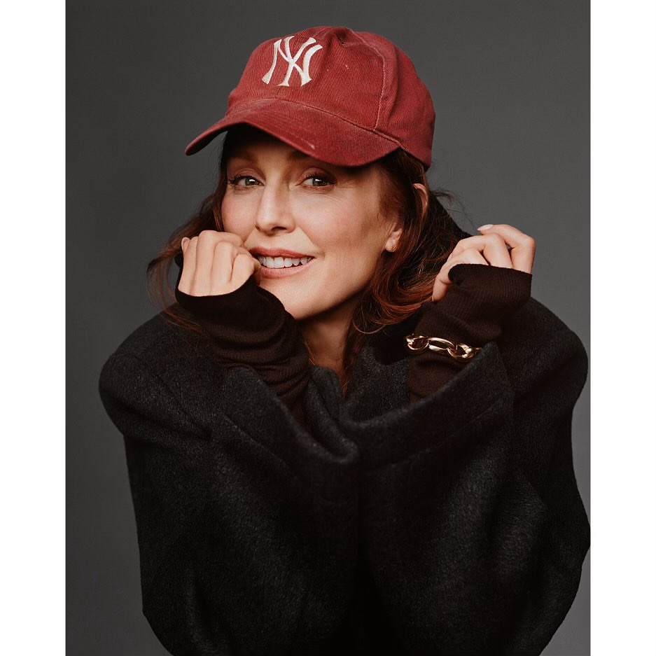 Julianne Moore smiling while wearing a red baseball cap and dark oversized coat against a grey background