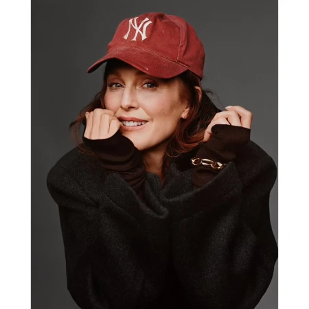 Julianne Moore smiling while wearing a red baseball cap and dark oversized coat against a grey background