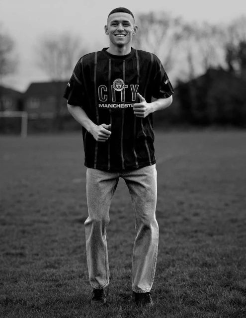 Monochrome portrait of a young adult male smiling on a grassy field wearing a striped jersey, jeans, and sneakers in an outdoor setting.