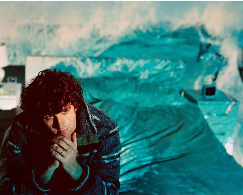Person with curly hair sits in a blue-toned bedroom with a bed and nightstand; their hands are clasped near their face.