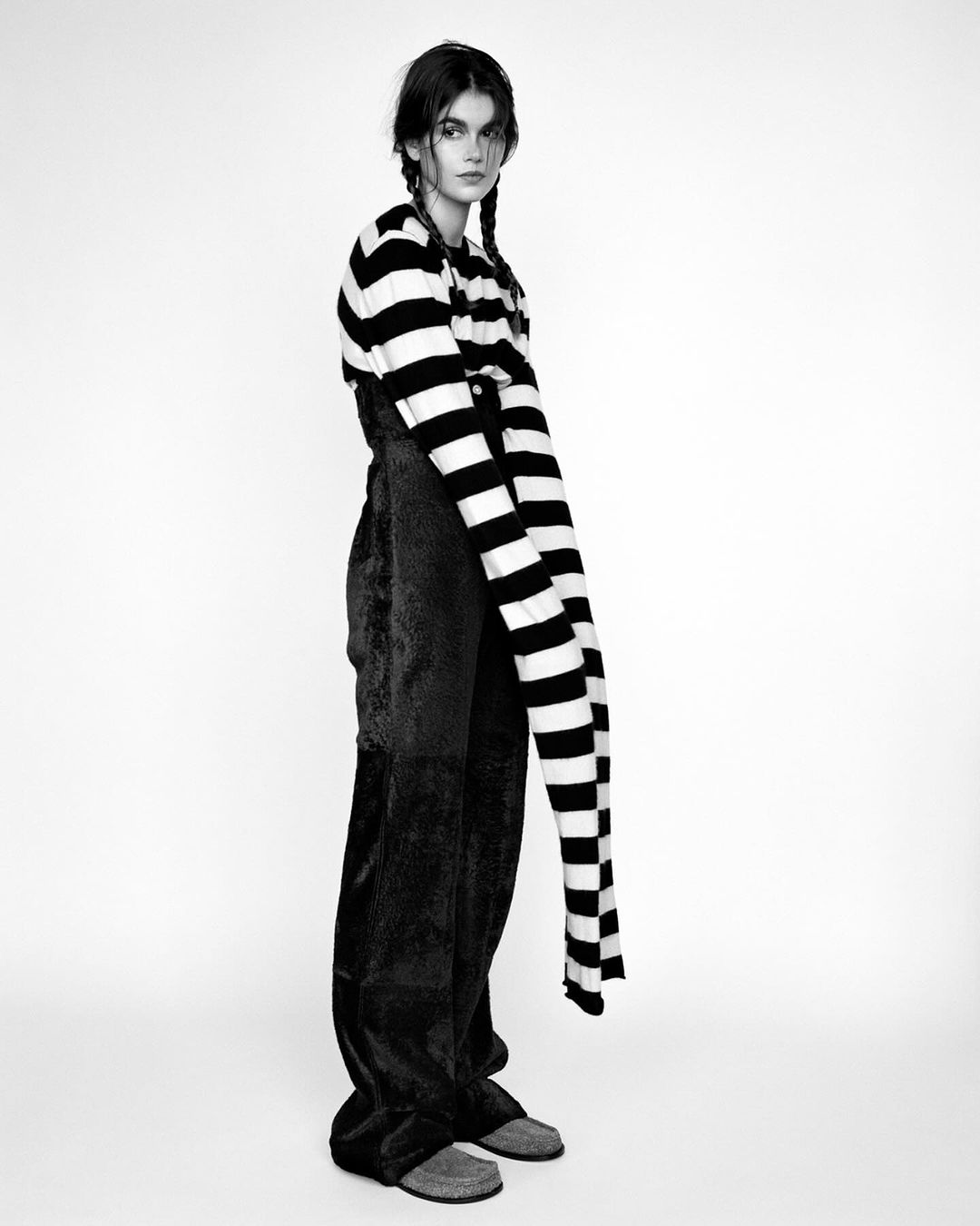 Monochrome portrait of a woman with braided hair wearing a striped sweater and dark pants, posing against a plain white backdrop in studio.