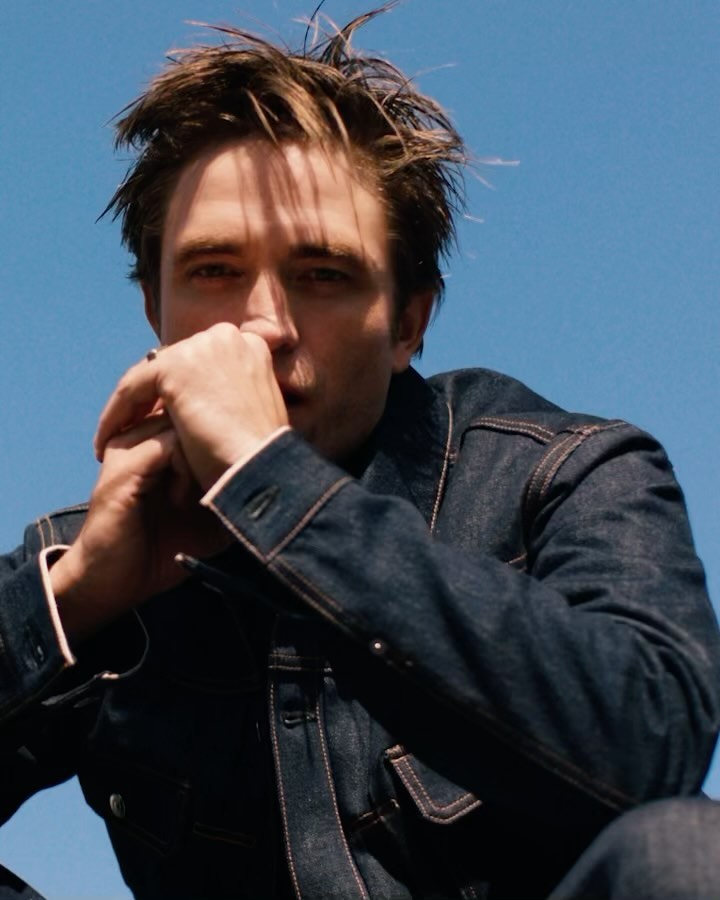 Robert Pattinson in denim jacket poses against a blue sky. He's looking at the camera with hands near his face in portrait shot.