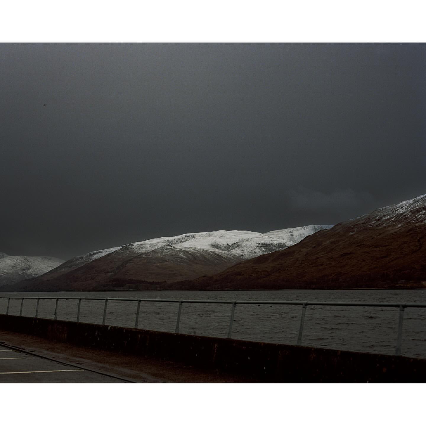 Snow-covered mountains are reflected in the still waters of a loch under a cloudy sky, framed by a concrete railing.