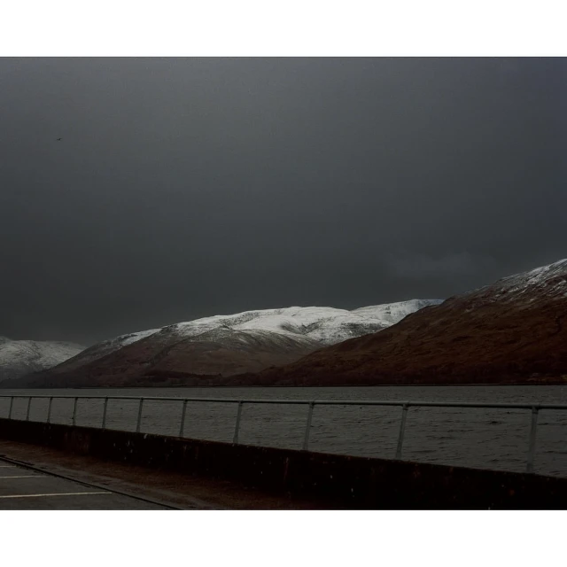 Snow-covered mountains are reflected in the still waters of a loch under a cloudy sky, framed by a concrete railing.