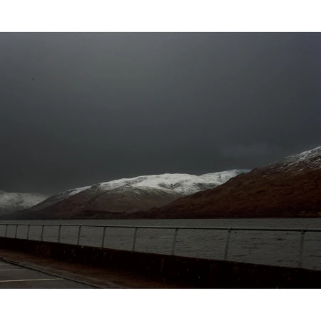 Snow-covered mountains are reflected in the still waters of a loch under a cloudy sky, framed by a concrete railing.