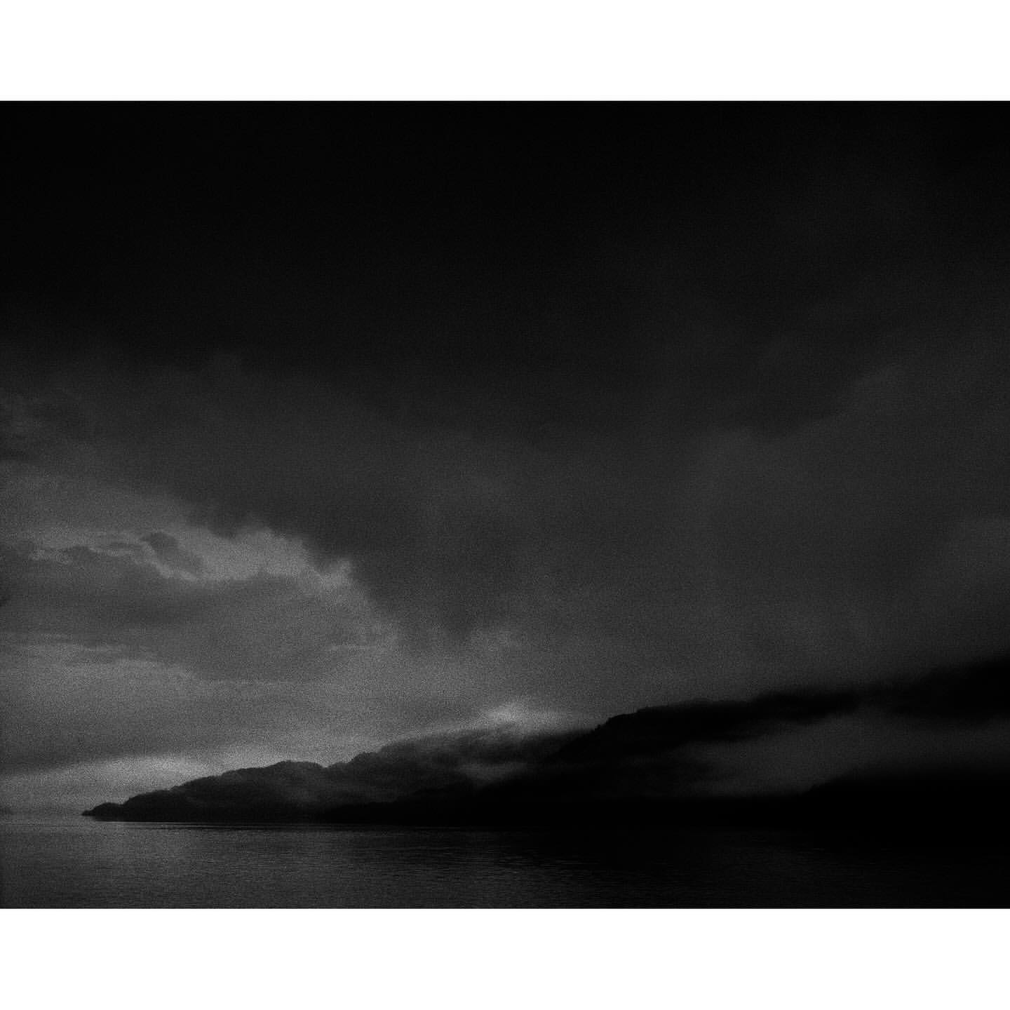 Monochrome landscape featuring a body of water next to a landmass covered in fog under a dramatic cloudscape with a dark sky.