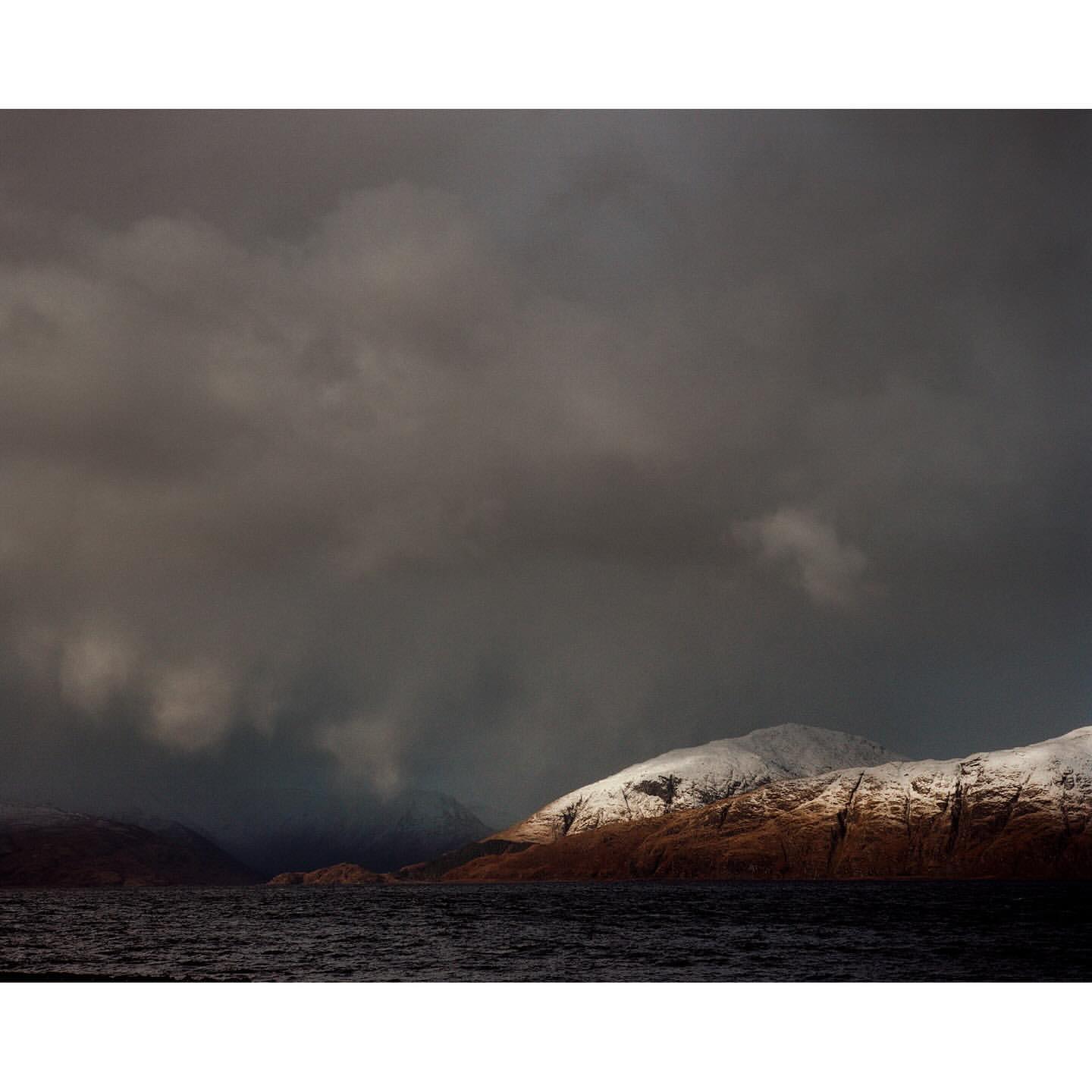 Gloomy seascape featuring a tranquil, dark blue sea contrasted by snow-covered mountains against an overcast, stormy sky with low clouds.