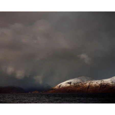 Gloomy seascape featuring a tranquil, dark blue sea contrasted by snow-covered mountains against an overcast, stormy sky with low clouds.