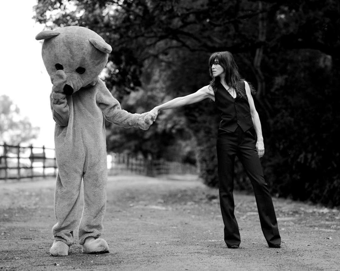 A woman in a vest and trousers holds hands with a person in a teddy bear costume on a path surrounded by trees in a black and white photograph.