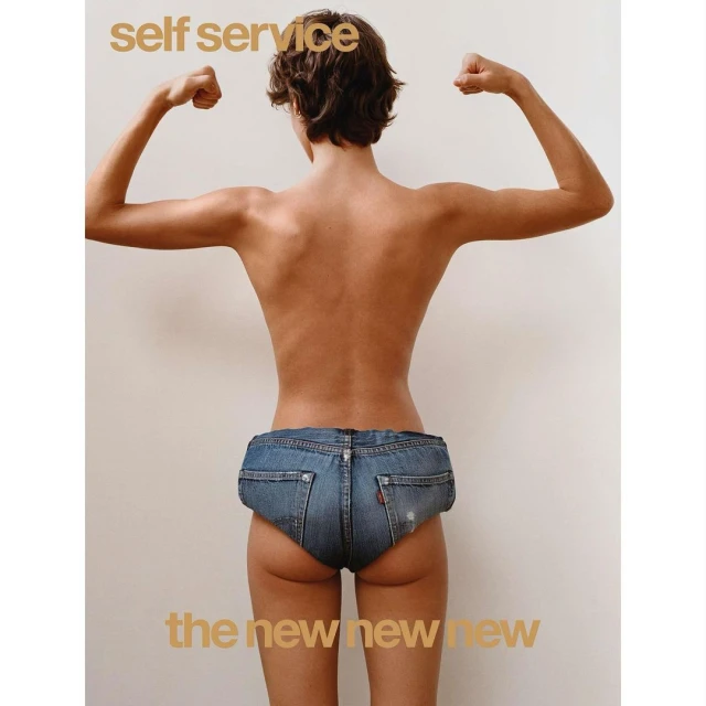 A slender adult woman faces away, flexing arm muscles, wearing denim shorts against a plain backdrop in a minimalist fashion editorial.