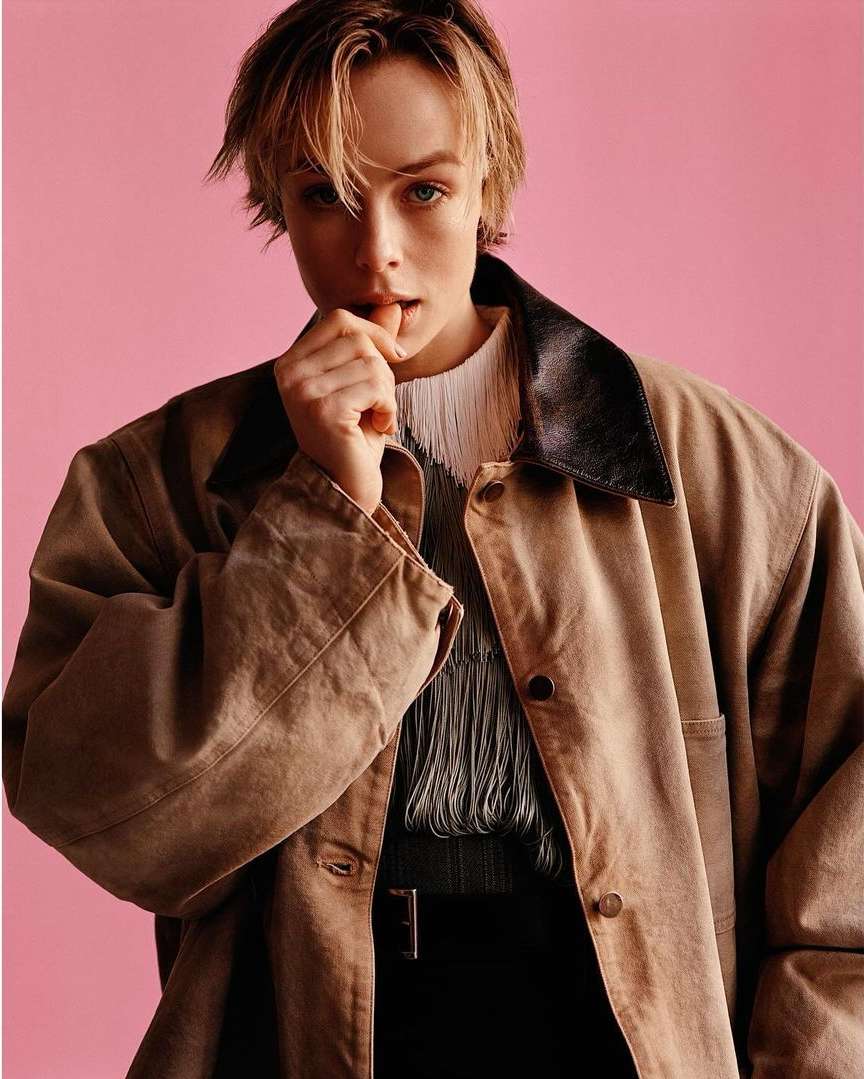 Portrait of a blonde female model with short hair, wearing a tan coat, and black pants against a pink backdrop in a photo studio.