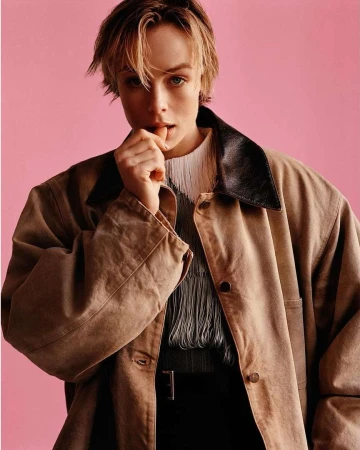Portrait of a blonde female model with short hair, wearing a tan coat, and black pants against a pink backdrop in a photo studio.