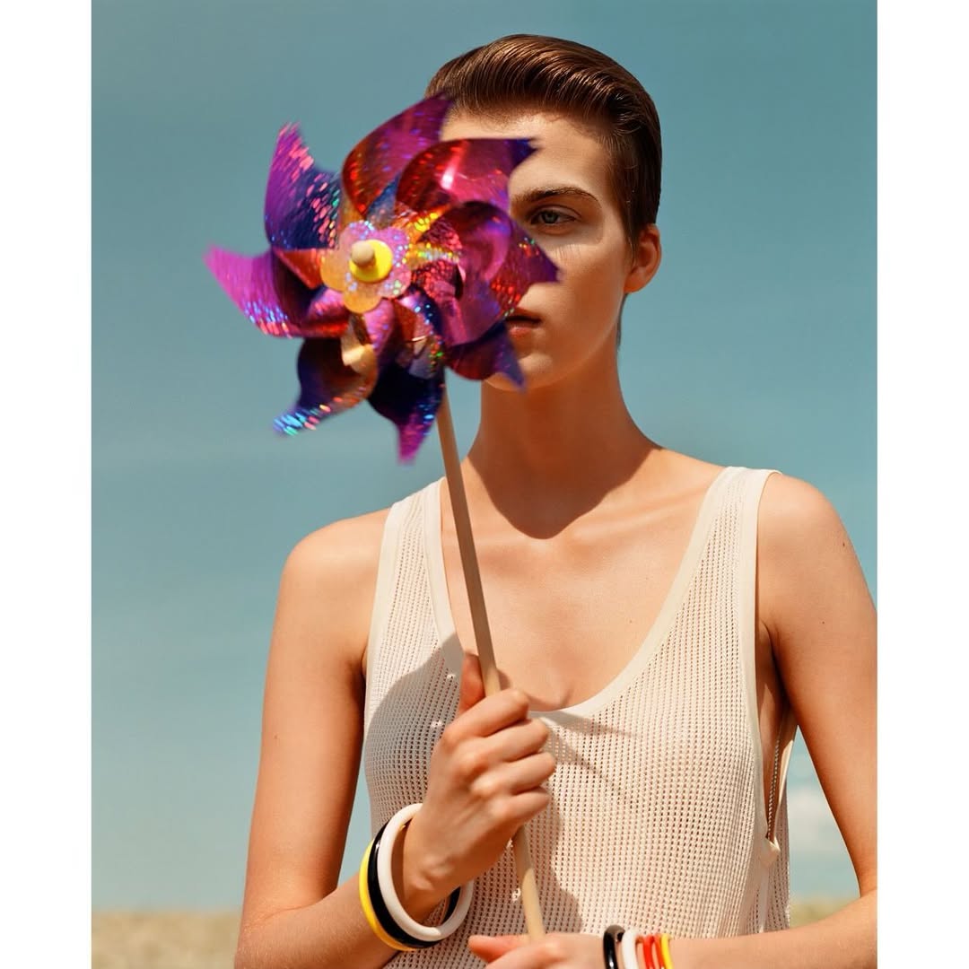 Young adult woman in a tank top poses outdoors holding a colorful pinwheel toy. She wears multiple bracelets. The background is blue sky.