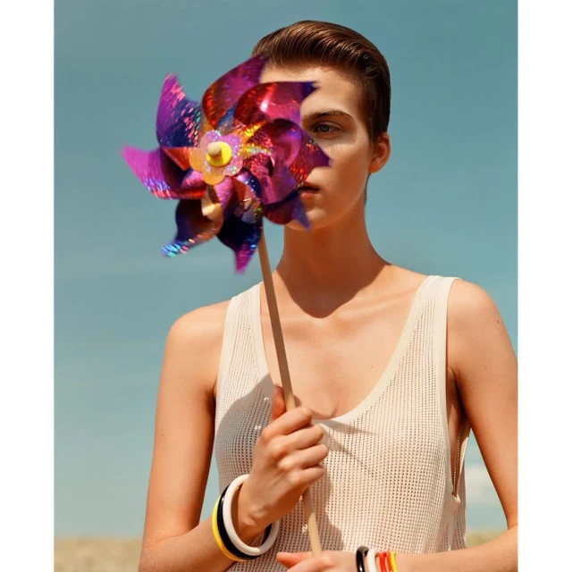 Young adult woman in a tank top poses outdoors holding a colorful pinwheel toy. She wears multiple bracelets. The background is blue sky.