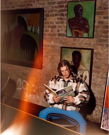 Young man in patterned fleece jacket and blue trousers seated indoors reading a book beneath framed art on a brick wall