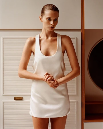 Model with slicked-back hair wearing a white scoop-neck slip dress against a paneled cabinet wall interior