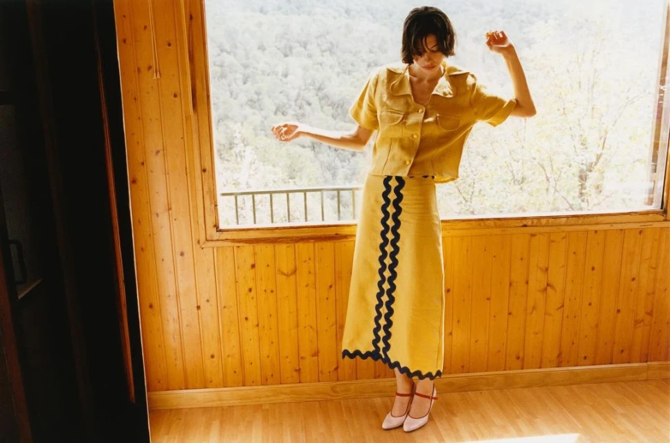 A woman models a yellow skirt and shirt in an indoor setting with a window view of trees; light catches the wooden floors.