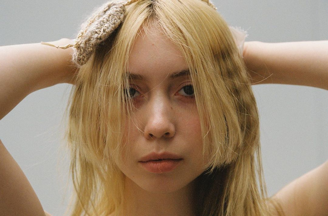 Close-up portrait of a fair-skinned young woman with long blonde hair, posing with her hands behind her head in a neutral studio setting.