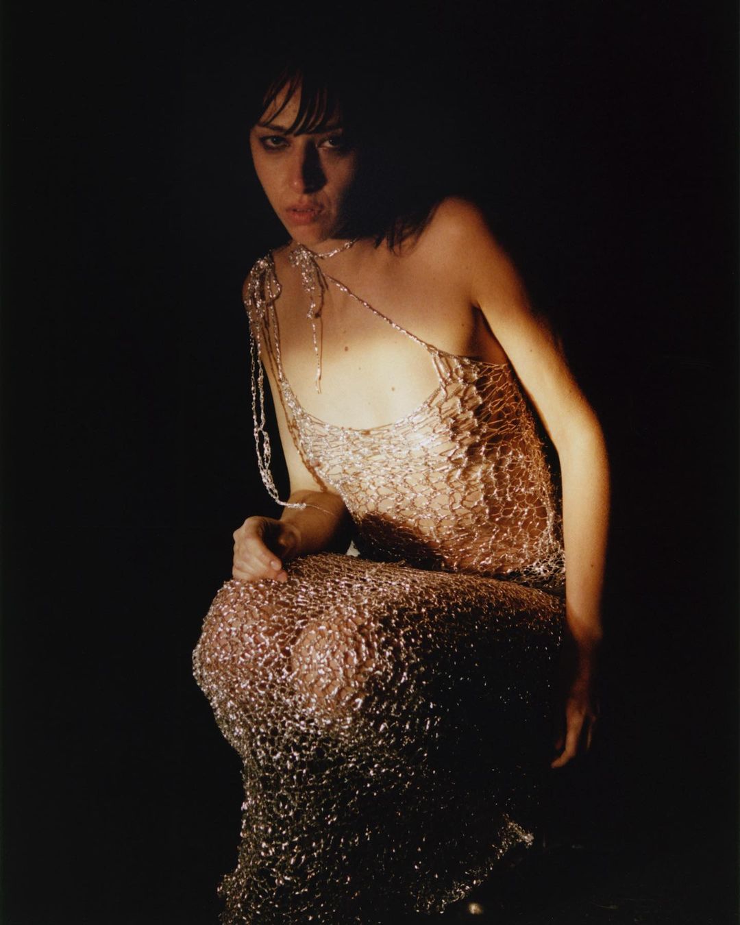 Adult female in textured shimmering one-shoulder dress poses in a dimly lit studio, enhancing the dress's unique and shiny design.