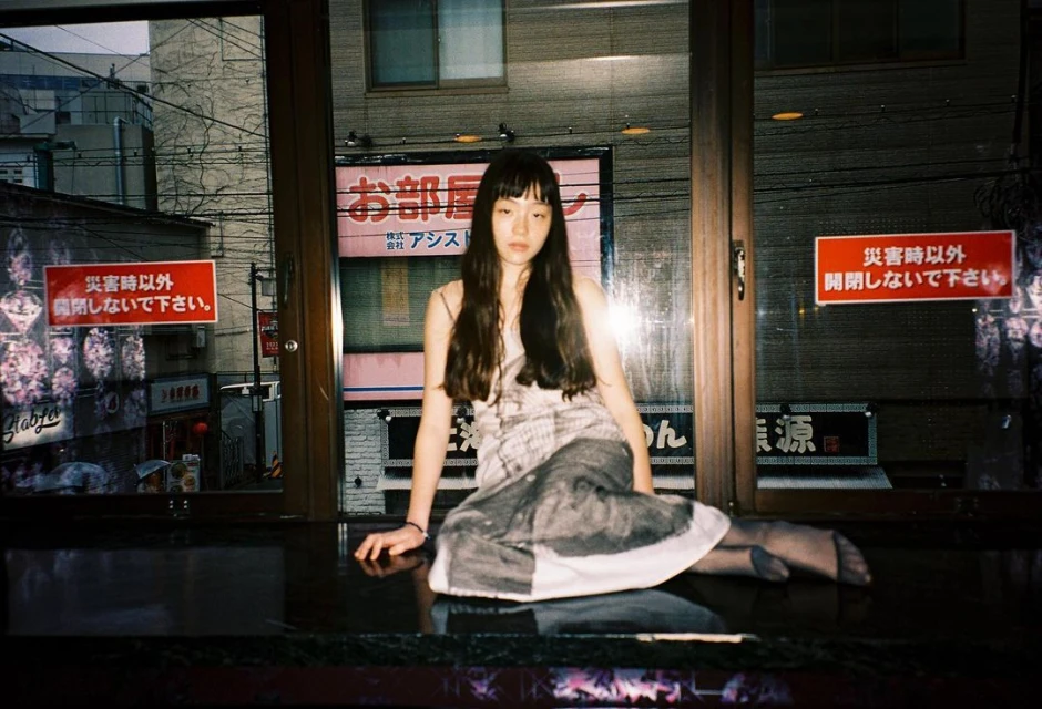 Young woman sits in an urban setting at night; she has long dark hair and is wearing a patterned dress and dark tights. 