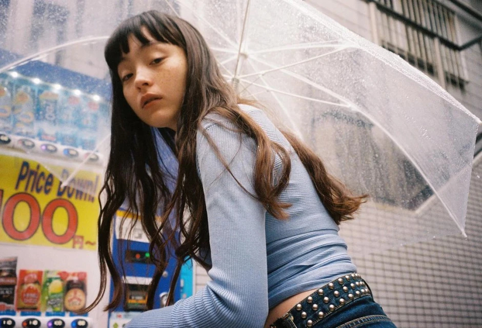 Young adult woman with long brown hair stands under a clear umbrella in an urban setting near a vending machine.