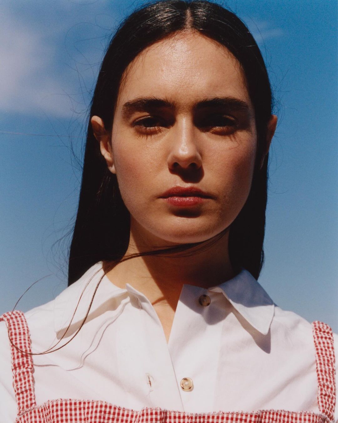 Close-up portrait of a young woman with long dark hair, wearing a white shirt with a red and white gingham pattern against a blue sky.