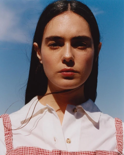Close-up portrait of a young woman with long dark hair, wearing a white shirt with a red and white gingham pattern against a blue sky.