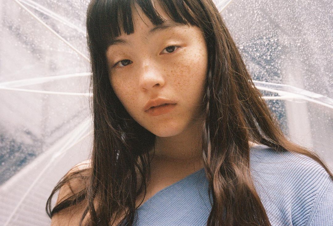 Close-up portrait of a young adult woman with freckles and long brown hair, wearing a blue top, with a clear umbrella background.