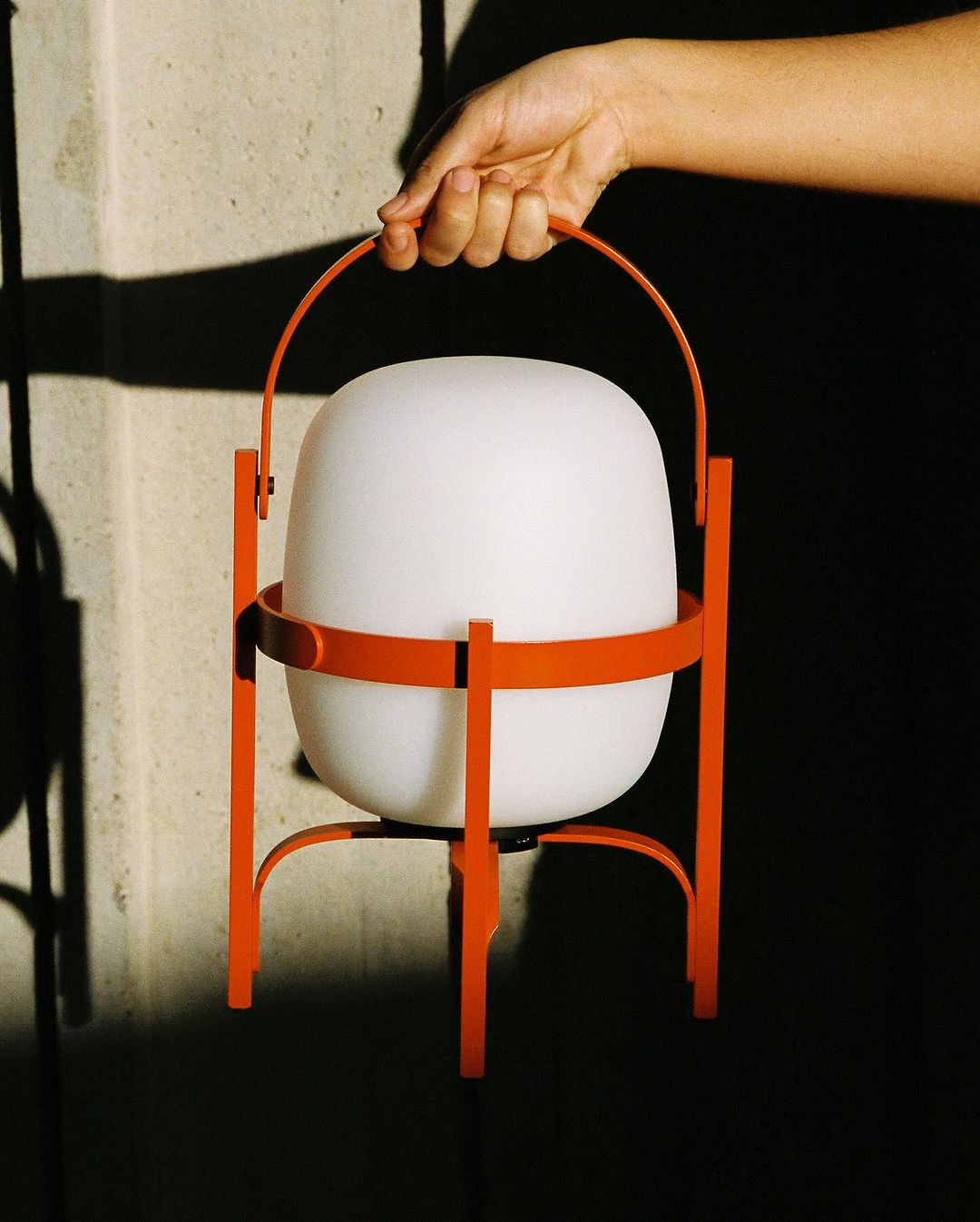 A hand holds a modern, portable lamp with a glowing white shade and an orange metal frame, against a neutral wall with shadows.