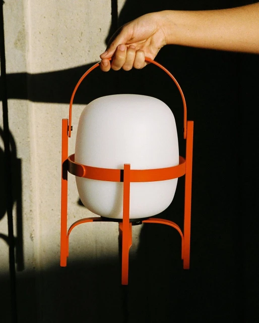 A hand holds a modern, portable lamp with a glowing white shade and an orange metal frame, against a neutral wall with shadows.