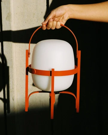 A hand holds a modern, portable lamp with a glowing white shade and an orange metal frame, against a neutral wall with shadows.
