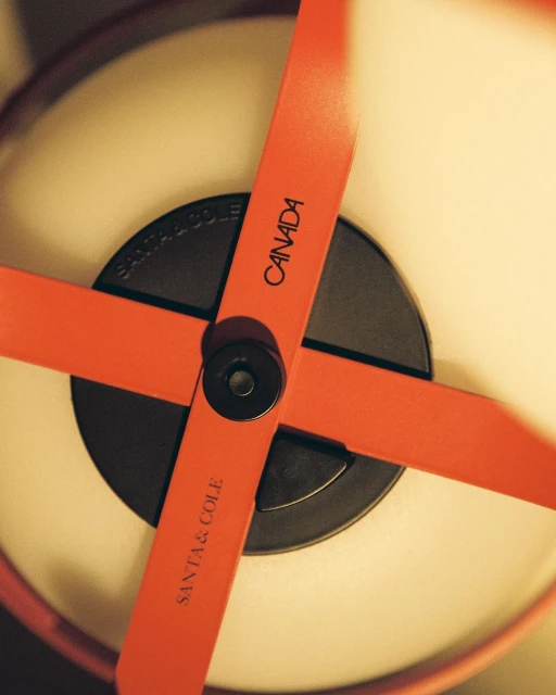 Close-up shot of a modern lamp featuring a black cylindrical base with orange crossbars, displaying the text "SANTA & COLE CANADA".