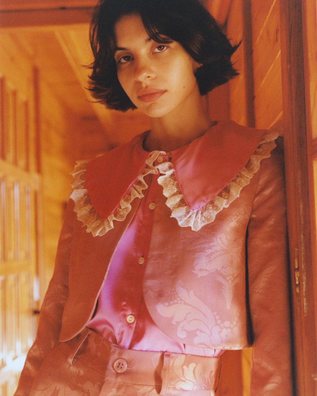 Young woman in pink brocade jacket and pants poses indoors with a ruffled collar. Fashion, style, and portrait photography.