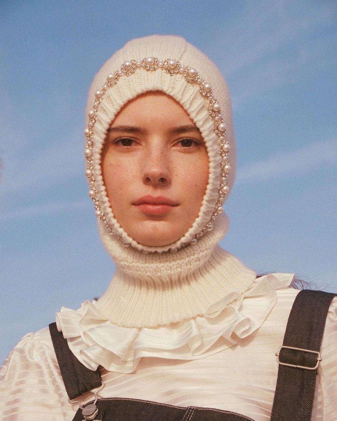 Close-up of a woman with a neutral expression wearing a white, pearl-trimmed balaclava, a frilled top, and dark denim overalls against a blue sky.