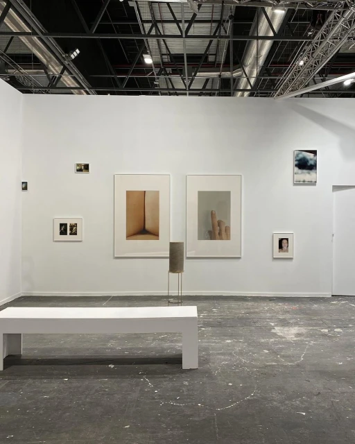 Gallery featuring framed art on white walls, a minimalist bench, and an industrial ceiling with metal beams and ductwork. Modern, clean design.