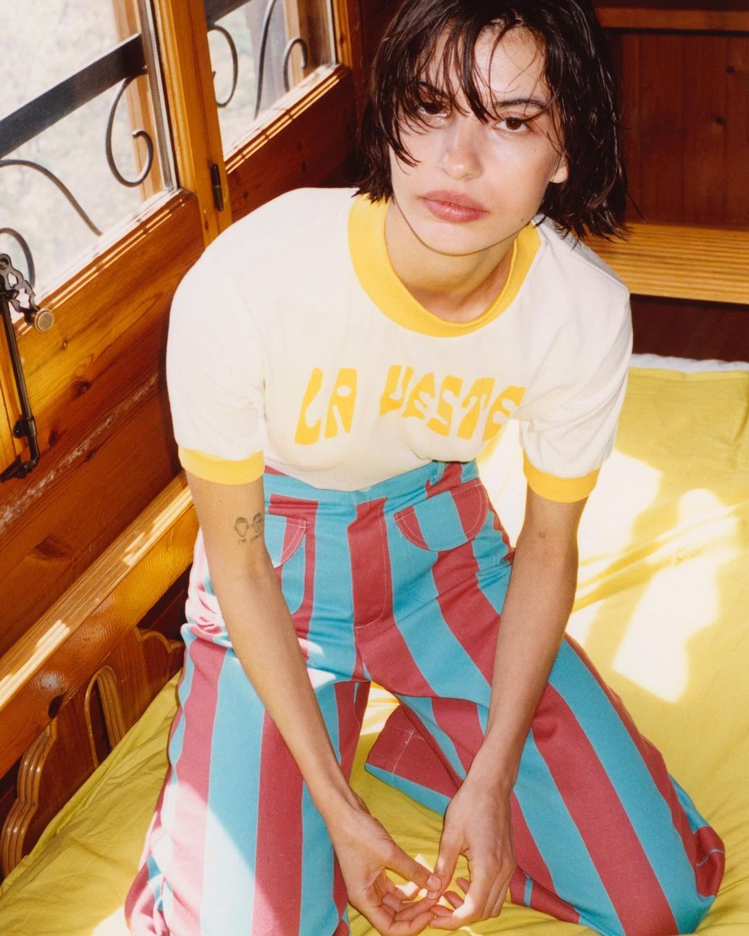 Young woman with short dark hair poses indoors wearing a white tee shirt and striped pants, seated on a yellow mattress.