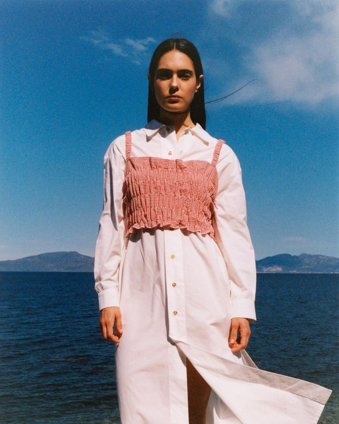 Adult female model wears pink crop top and white dress against a blue sky and ocean backdrop. Fashion shot outdoors in daylight.