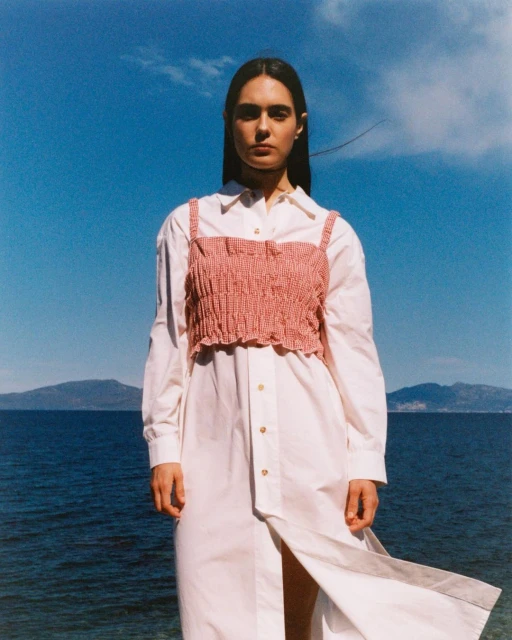 Adult female model wears pink crop top and white dress against a blue sky and ocean backdrop. Fashion shot outdoors in daylight.