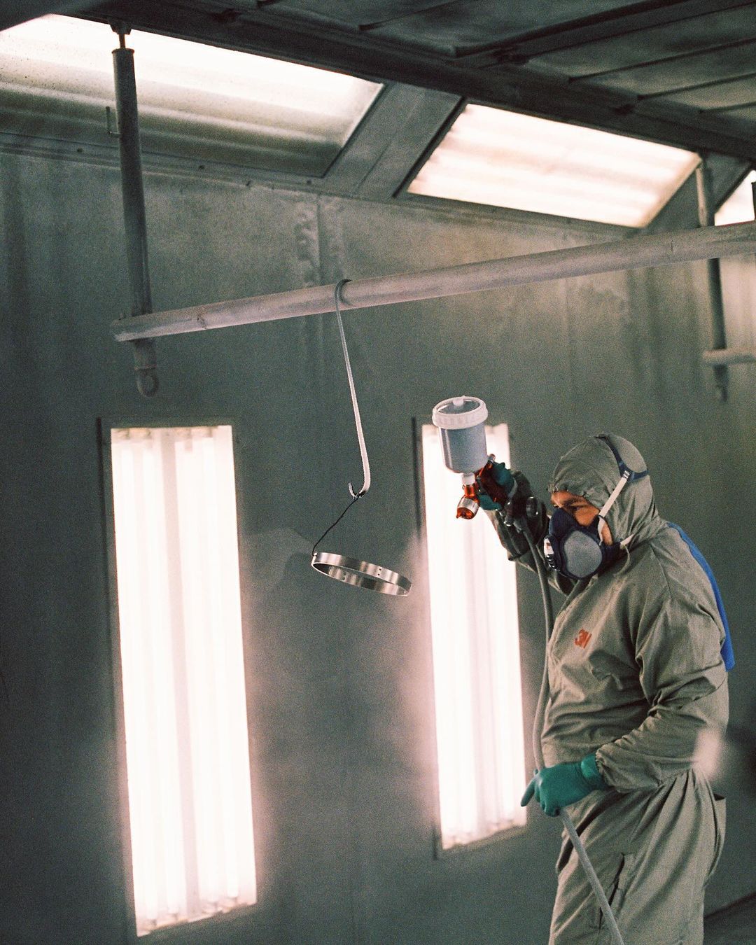 Adult male wearing protective gear spray paints metal objects in a paint booth with fluorescent lighting. Grey color palette dominates the image.