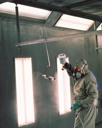 Adult male wearing protective gear spray paints metal objects in a paint booth with fluorescent lighting. Grey color palette dominates the image.