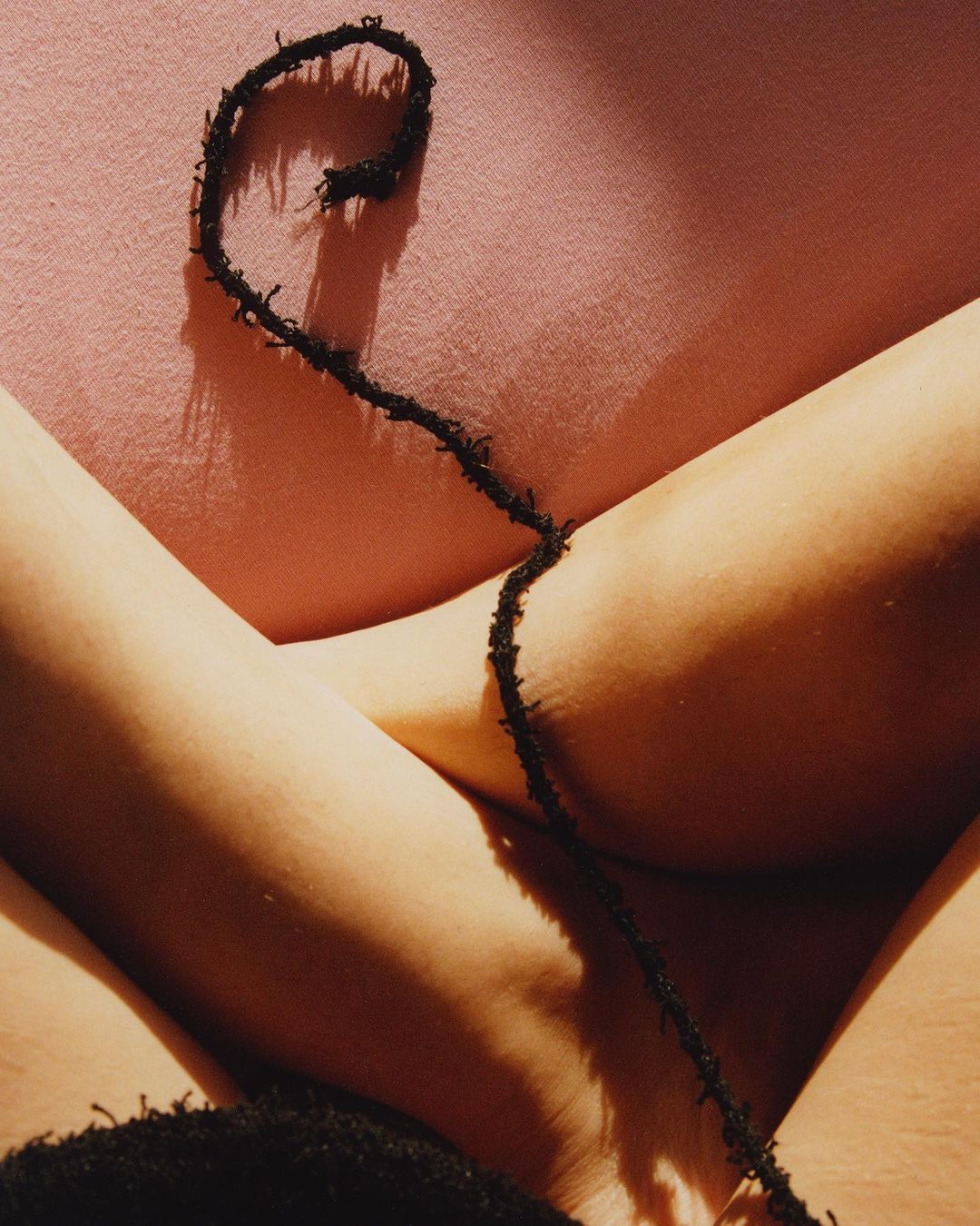 Close-up shot of a person's crossed legs and black string over a pink surface in a studio with bright, diffused lighting.