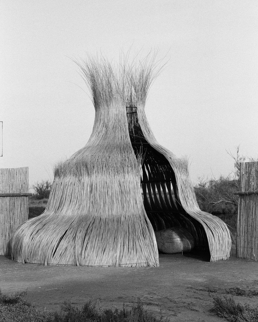 Unique straw structure in a monochromatic desert landscape. The building has a bulbous base, narrow midsection, and a flared top.