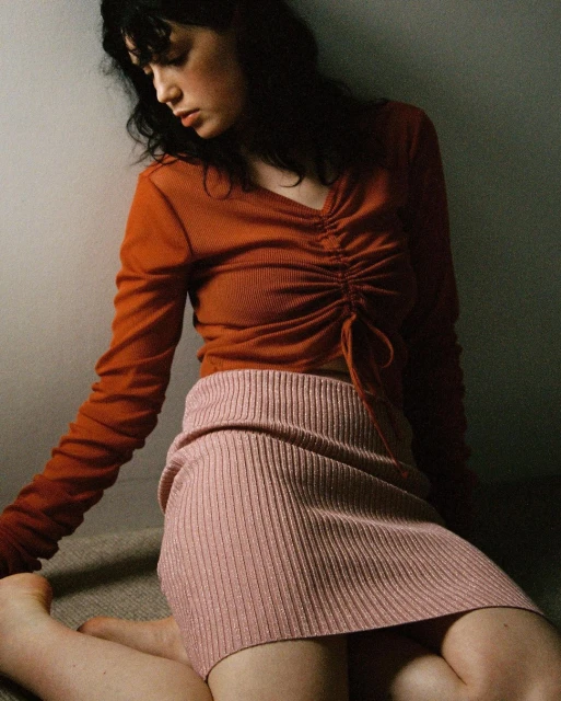 A young woman kneels indoors wearing a rust-colored long-sleeve top and a pink ribbed skirt, looking down with a neutral expression.
