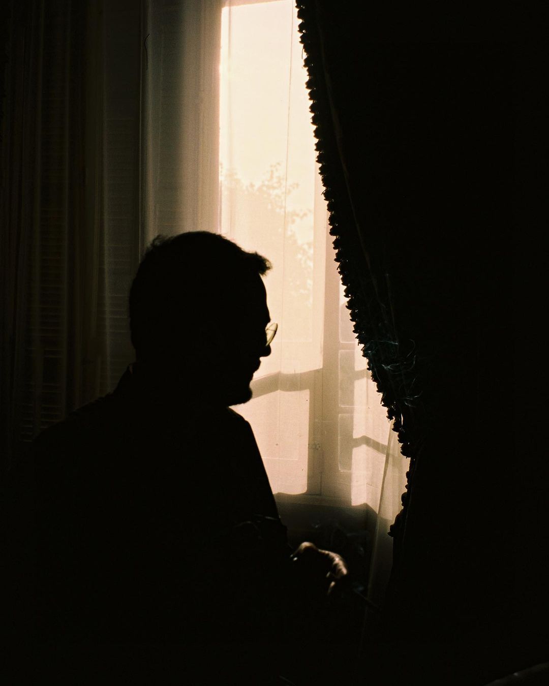 Silhouette of adult male with glasses near a window with curtains creates contrast and mystery in dimly lit, evocative indoor setting.