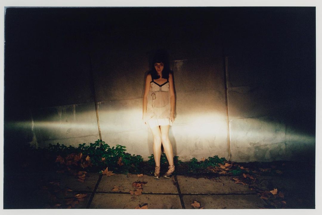 Full-length portrait of a woman in a light dress standing against a concrete wall in the dark, illuminated by car headlights.