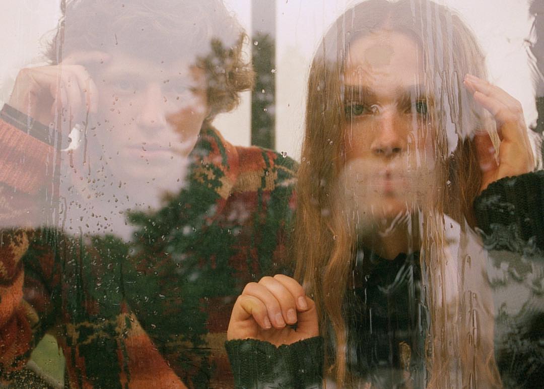 Couple looking through a rain-streaked window. The woman is in the foreground, with her hand resting on the glass, and the man is behind her.